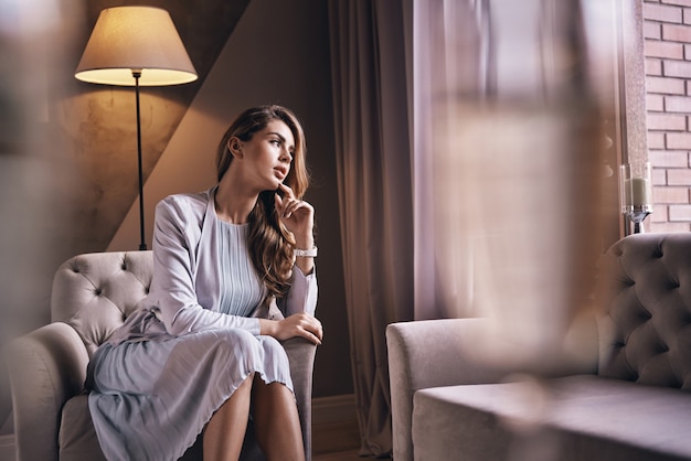 What is she thinking about? Attractive young woman keeping hand on chin and looking away while sitting in the armchair