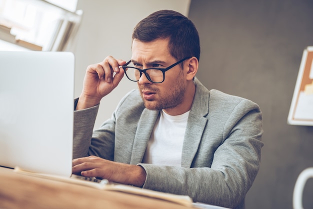 What is this? Handsome young man looking at his laptop and adjusting his glasses while sitting at his working place