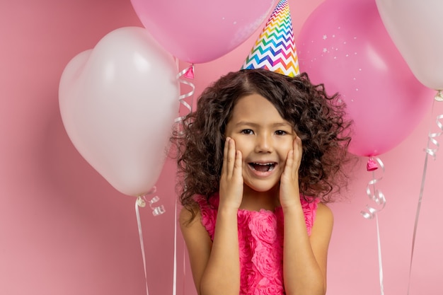 What a surprise. Cute overjoyed caucasian little girl in dress, party hat, standing among balloons with open mouth, keeping palms on checks, receiving long awaited gift, posing.