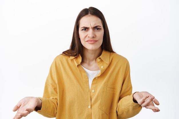 Photo whats wrong i dont understand, confused woman frowning and raising hands clueless, cant tell, know nothing, why bother gesture, standing over white background