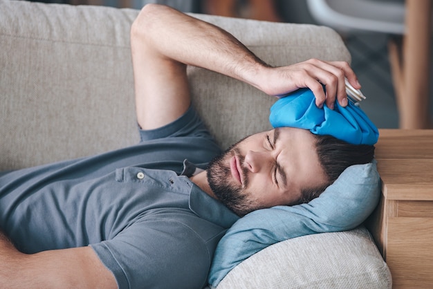 When you hit shelf with your head. Frustrated young man holding ice bag on his head while lying on the couch at home