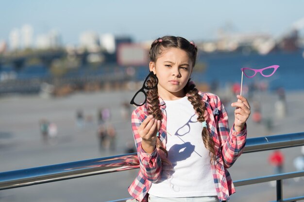 Which props to choose Doubting small girl choosing glasses props Adorable little child holding photobooth props on sticks Cute kid with fancy party props on urban outdoor