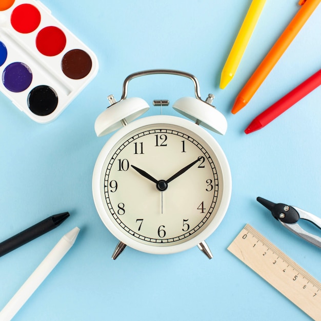 White alarm clock surrounded by colored pens and other school supplies. Art concept of the beginning of the school year