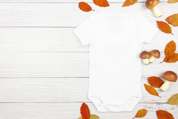 White baby girl or boy bodysuit mockup flat lay with mushrooms and fallen leaves on wooden background Design onesie template print presentation mock up Top view