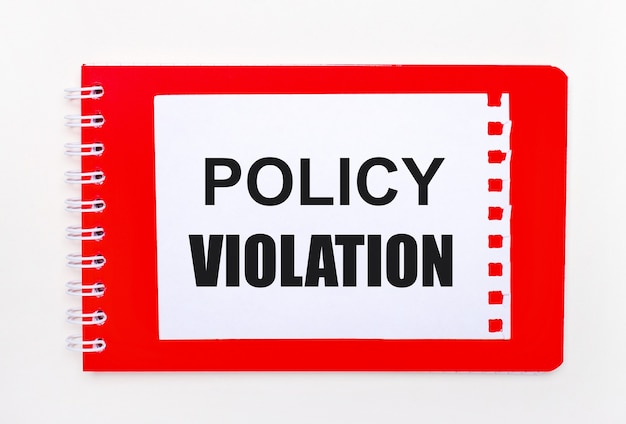 On a white background - a bright red notebook on a spiral. On it is a white sheet of paper with the text POLICY VIOLATION