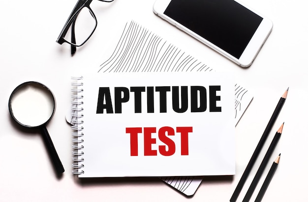 On a white background glasses, a magnifier, pencils, a smartphone and a notebook with the text APTITUDE TEST