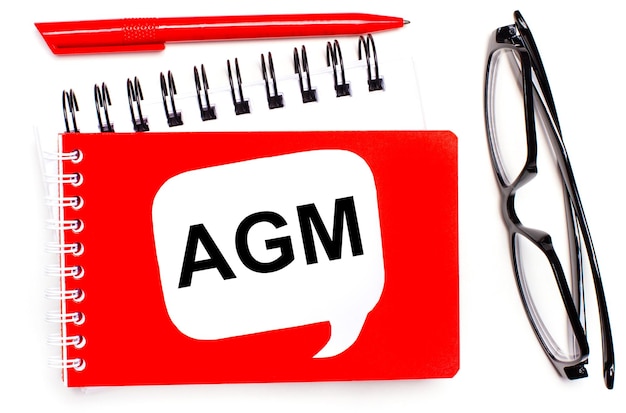 On a white background white and red notepads black glasses a red pen and a white card with the text AGM Annual General Meeting