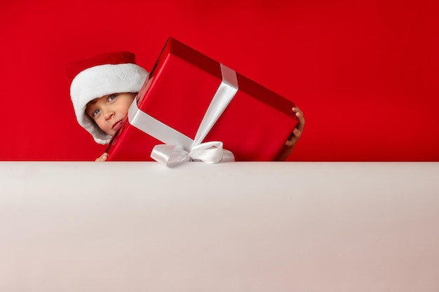 Next to a white billboard A beautiful Christmas boy holds a red gift in studio