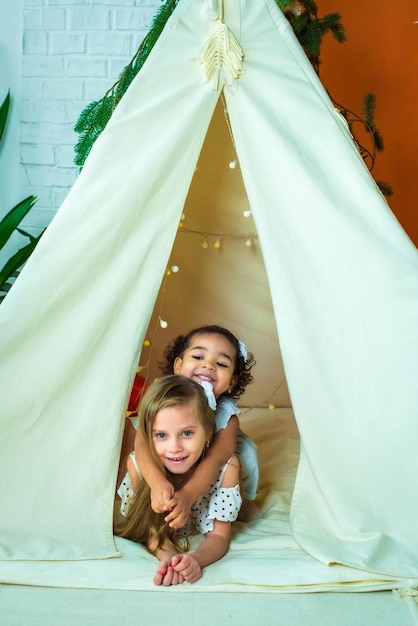 White and black africanamerican girls play in a tent wigwam kids laugh have fun happy childhood