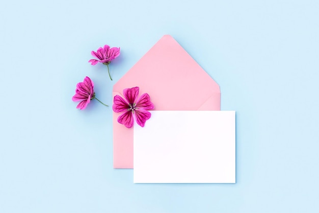 White blank card pink envelope and flowers on blue background Minimal style Top view Flat lay Mockup