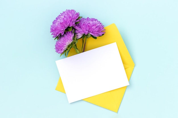 White blank card yellow envelope and purple flower on blue background Minimal style Top view Flat lay Mockup