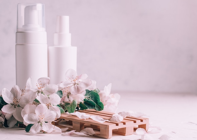 White bottles with cosmetic products on wooden podium in form of pallet on light plaster surface with apple flowers. Copy space