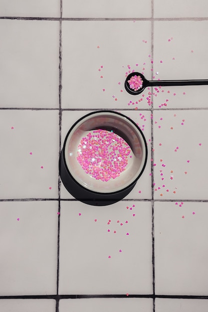 A white bowl and a black teaspoon with pink hearts glitter sequins on a white square tile counter.