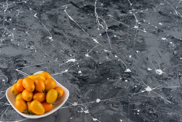 A white bowl full of fresh cumquat fruits placed on a marble table .