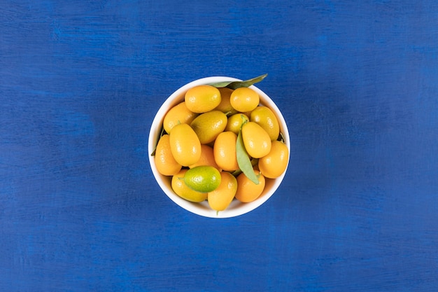 White bowl full of yellow kumquats on blue surface.