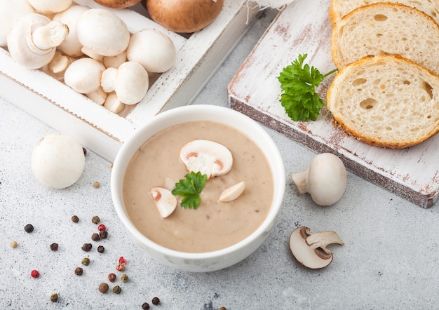 White bowl plate of creamy chestnut champignon mushroom soup on light kitchen background and box of raw mushrooms and fresh bread. Top view