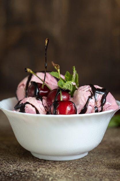 White bowl with pink ice cream balls, cherry berries and mint on the ice cream poured with chocolate, brown wooden background, morning dessert, vertical