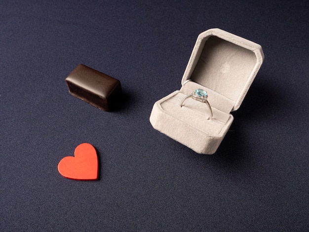 White box in which a ring with a blue stone, a red heart and chocolate lies nearby on a blue