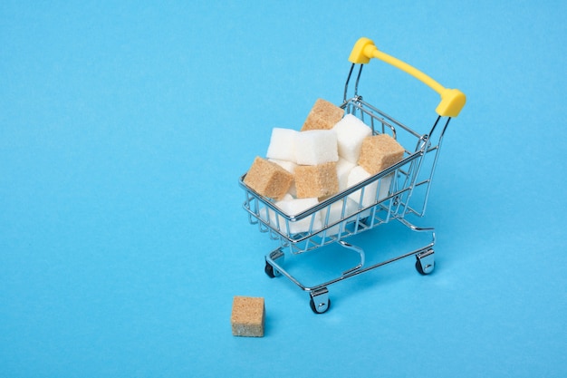 White and brown sugar cubes in a shopping cart on a blue surface copy space