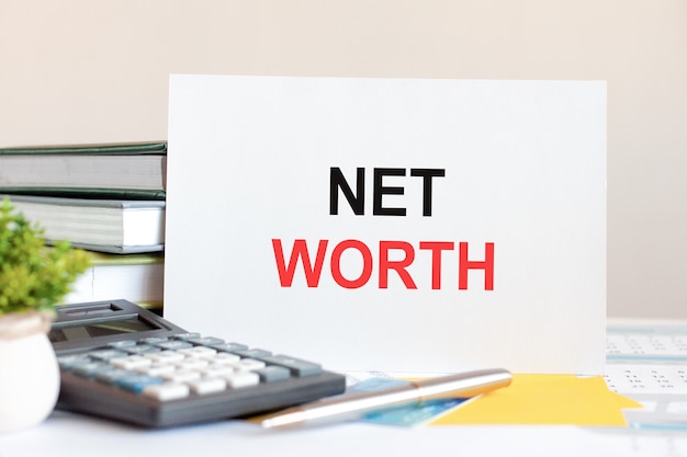 White card with text net worth stands on the desk against the background of books stacked, calculator, pen, green potted plant. Business and financial concept. Selective focus.