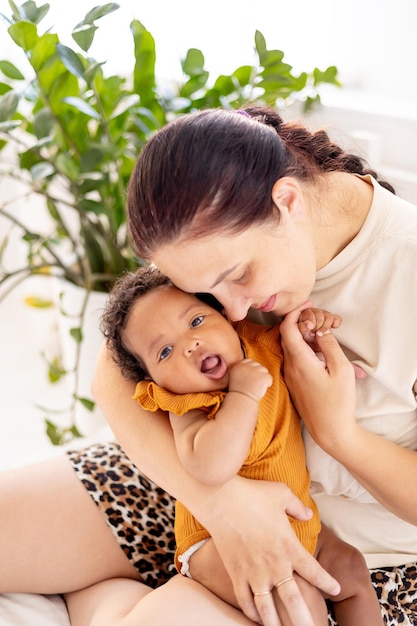 white Caucasian mom hugs and kisses an AfricanAmerican black foster baby girl at home on the bed maternal love and care a mother of European appearance with a black child in her arms