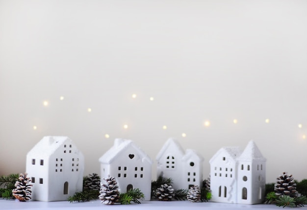 White ceramic houses with windows next to green spruce branches and snow-covered cones on a light blurred background with garland lights, horizontal image with soft focus and place for text