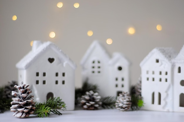 White ceramic houses with windows next to green spruce branches and snow-covered cones on a light blurred background with garland lights, horizontal image with soft focus and place for text