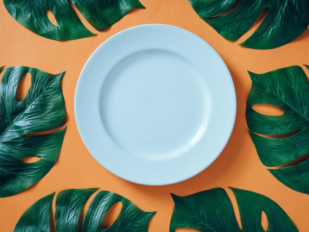 White ceramic plate with tropical leaves Monstera on orange background.