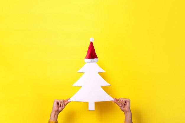 White Christmas tree in Christmas hat in hands over yellow background, merry christmas concept