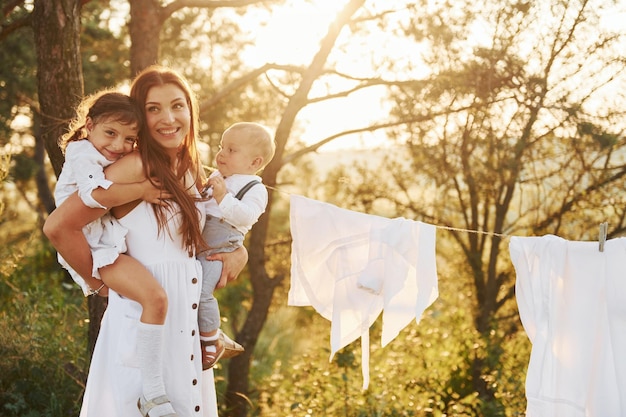 White clothes hanging on the rope to dry Young mother with her little daughter and son is outdoors in the forest Beautiful sunshine