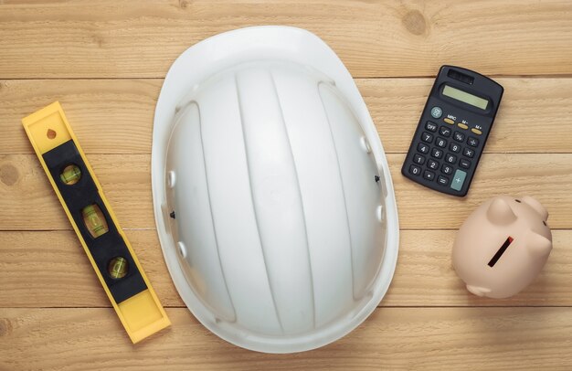 White construction safety helmet with level, piggy bank and calculator on wooden surface. The cost of repairing or building house. Top view.