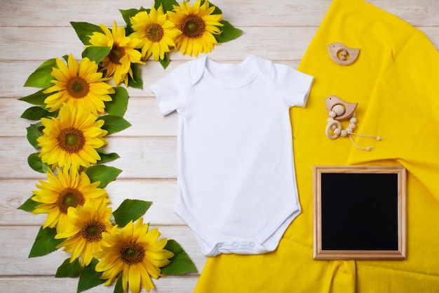 White cotton baby short sleeve onesie mockup with yellow sunflowers wooden children bird toys black chalkboard Design gender neutral bodysuit template newborn romper print presentation mock up