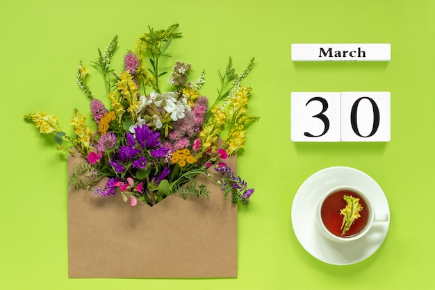 white cubes calendar March 30.Cup of tea, kraft envelope with multi colored flowers on green 