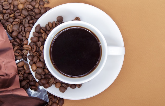 White cup of Americano black coffee without milk with a bunch of roasted coffee beans scattered from paper packaging. Coffee background, top view with copy space for logo or text.