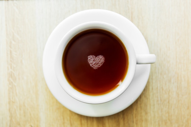 White cup of black tea with bubbles in a shape of heart. White cup on wooden table