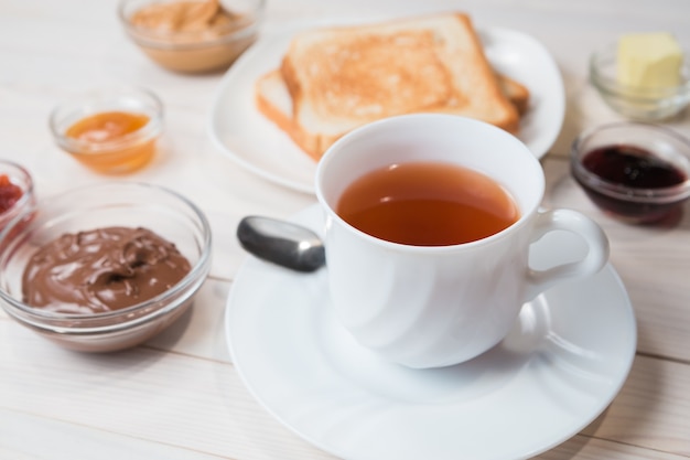 A white cup of black tea with Sandwiches or toasts with peanut butter, chocolate paste and strawberry, currant and apricot jelly or jam on white wooden table, close up, Breakfast concept