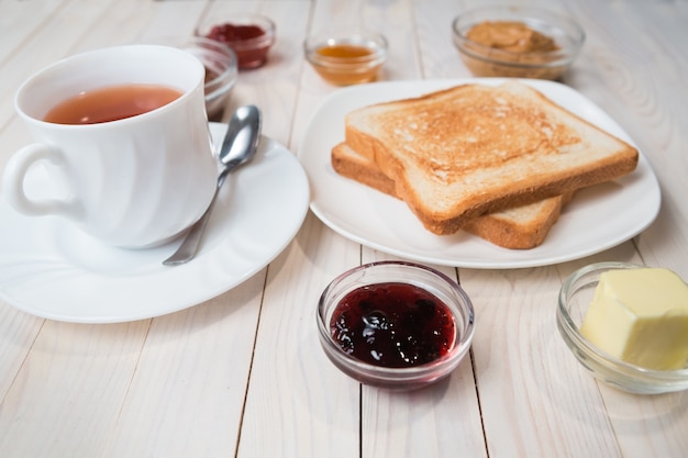 A white cup of black tea with Sandwiches or toasts with peanut butter, chocolate paste and strawberry, currant and apricot jelly or jam on white wooden table, close up, Breakfast concept