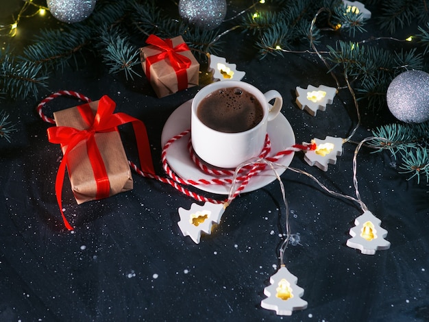 A white Cup of coffee, Christmas gifts, and a Christmas tree-shaped garland on a dark table in the evening. Homely holiday atmosphere