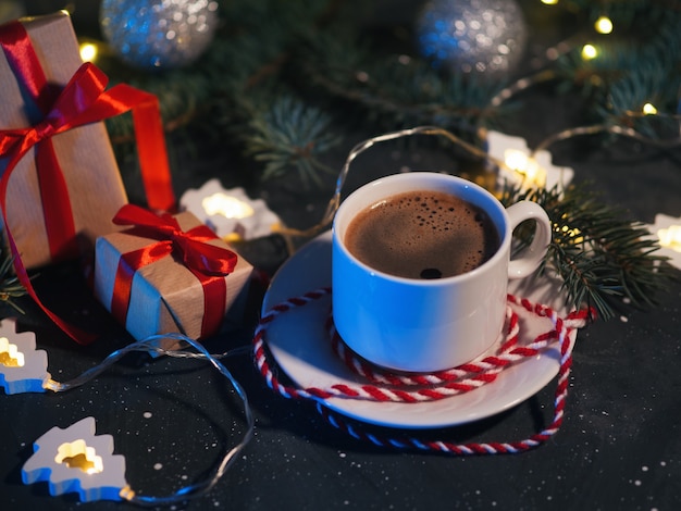 A white Cup of coffee, Christmas gifts, and a Christmas tree-shaped garland on a dark table in the evening. Homely holiday atmosphere