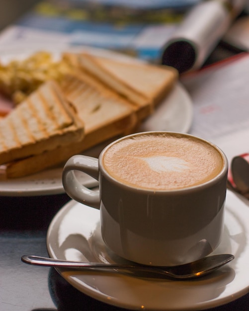 White cup of coffee in the foreground served on a breakfast table, in the background out of focus a breakfast of toast