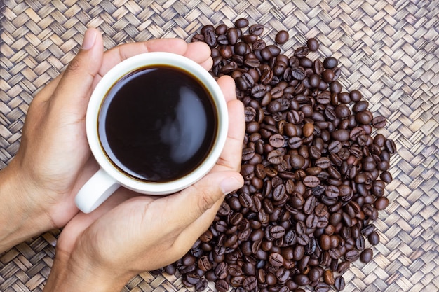 A white cup of coffee in hands of man with coffee beans next to him.