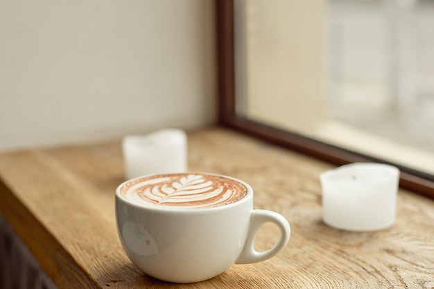 White cup of coffee with milk with a pattern on milk foam on a wooden windowsill