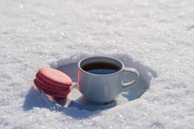 White cup of hot coffee with pink macaroon on a bed of snow and white background, close up. Concept of christmas winter morning