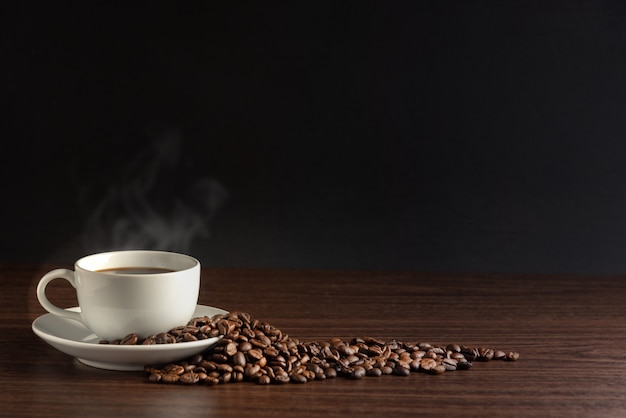 White cup of hot coffee with smoke with coffee beans and on black background