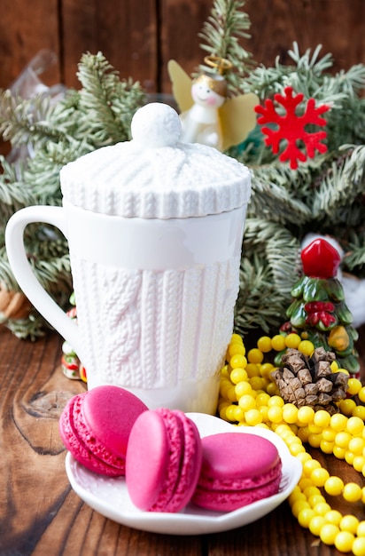 White cup and pink macaroons with christmas decorations on wooden background