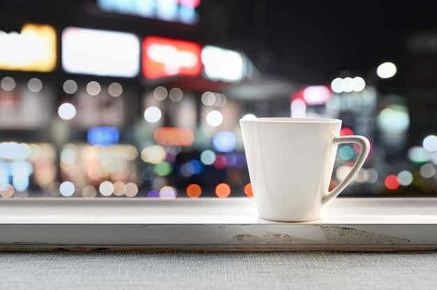 White cup placed on the window side with bokeh of night light background.