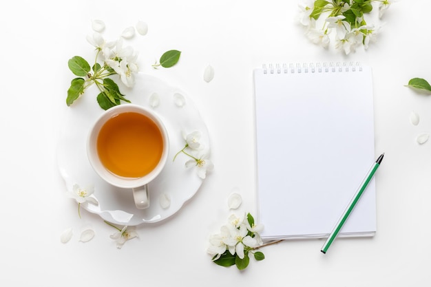 White cup of tea empty notebook with pen white apple tree flowers on white Tea time Top view