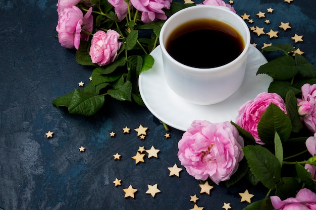 White cup with black coffee, stars and pink roses on a dark blue surface