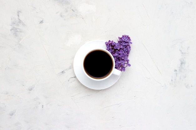 White cup with a coffee and beautiful lilac flowers on a white surface