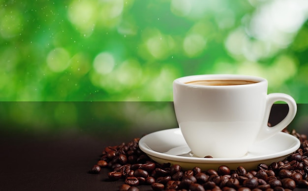 White cup with roasted coffee beans on black table against green background